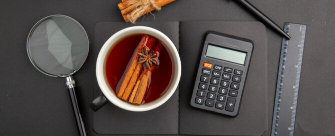 Tasse mit dunklem Gewürztee mit Zimtstangen und Sternanis; daneben Taschenrechner, Lineal, Stift und Lupe auf schwarzer Unterlage.