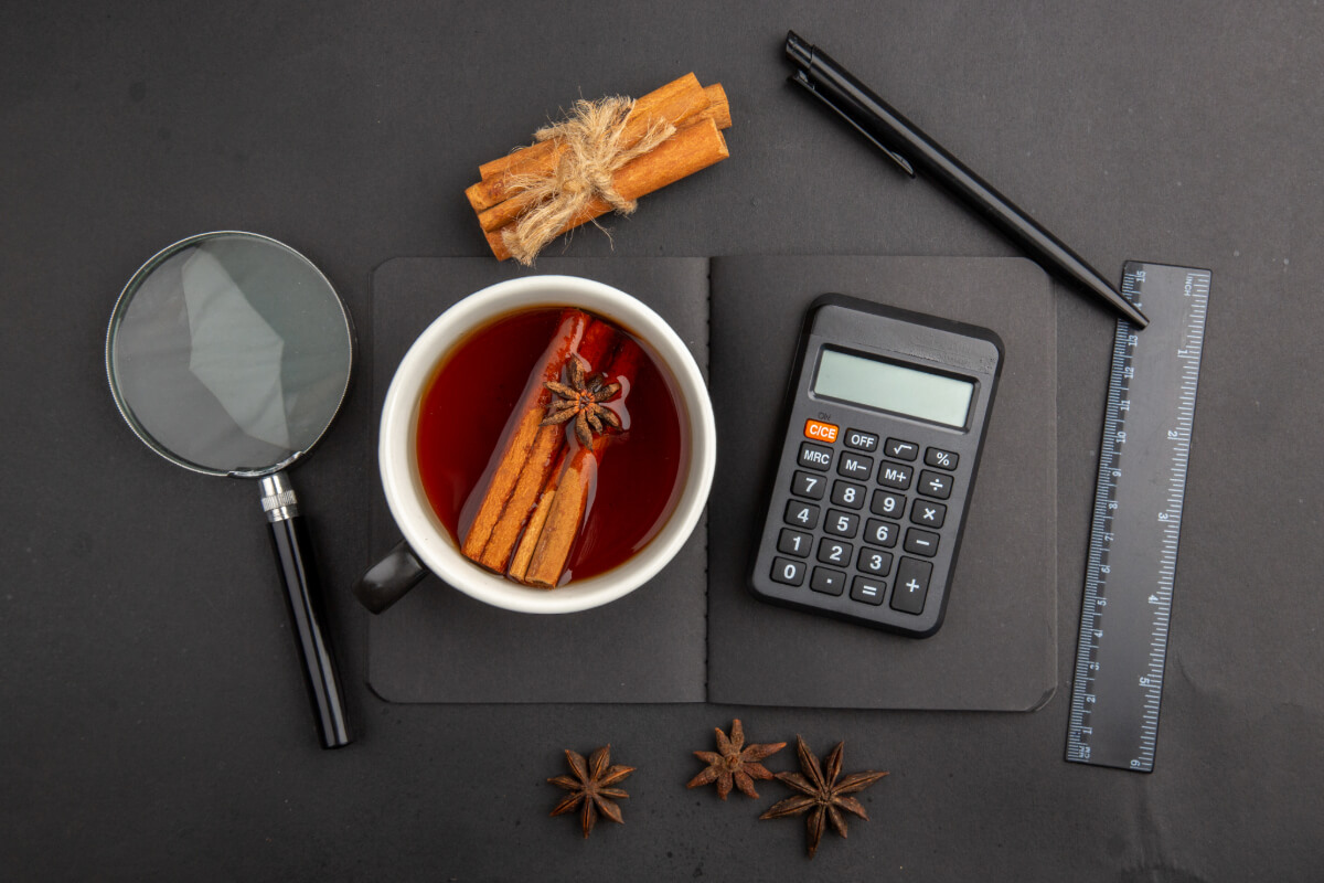 Tasse mit dunklem Gewürztee mit Zimtstangen und Sternanis; daneben Taschenrechner, Lineal, Stift und Lupe auf schwarzer Unterlage.
