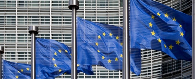 Mehrere Europaflaggen mit gelben Sternen auf blauem Grund wehen vor einem modernen Verwaltungsgebäude.